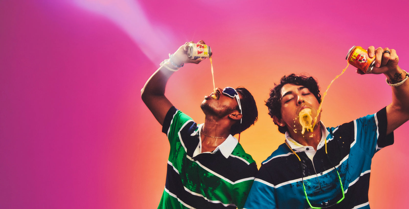 Two friends pouring cans of TIZZ tangerine soda into their mouths against a pink sky