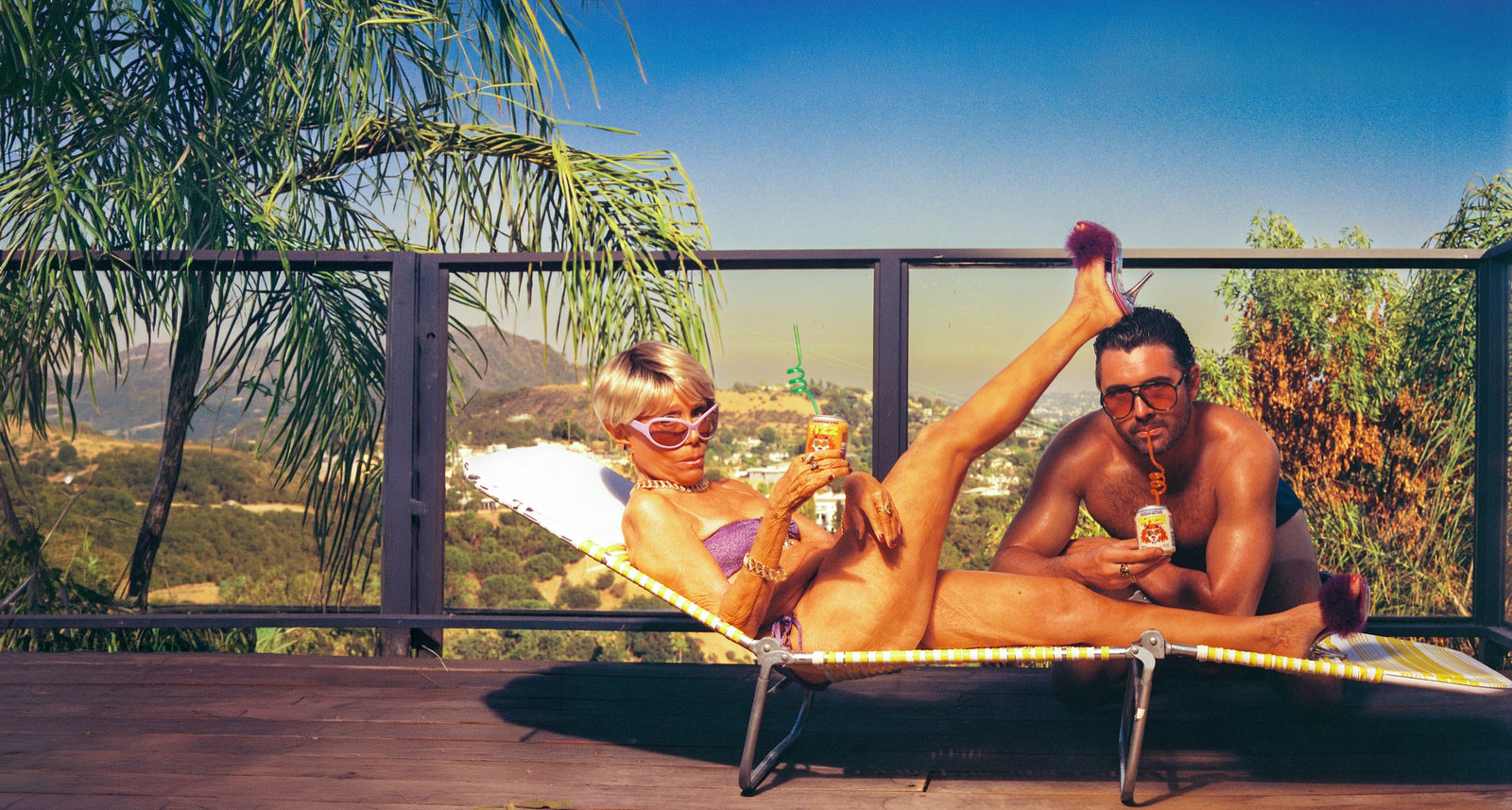 Couple lounging on a cot on a sun deck holding cans of TIZZ, palm trees and hills behind